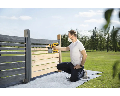 Muž natírá dřevěný plot stříkací pistolí na barvu na zahradě.
