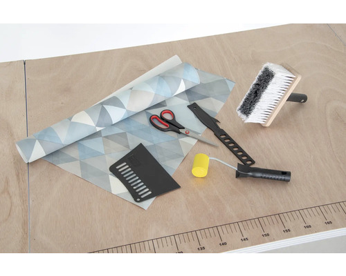 Tapeta s geometrickým vzorem, štětka na lepidlo, přítlačný váleček, tapetovací nůž, přítlačná špachtle a nůžky na stole.