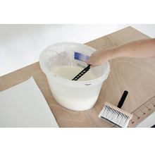 Lepidlo na tapety se míchá v kbelíku s míchadlem, vedle leží štětka na tapety a tapeta.