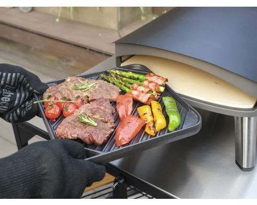 Osoba v kuchyňských rukavicích drží grilovací tác s grilovaným steakem, chřestem, rajčaty a paprikami před pecí na pizzu.