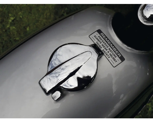 Detailní pohled na palivovou nádrž motocyklu s chromovaným uzávěrem a nálepkou připomínající ochranu přírody a bezpečnost