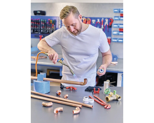 Muž pájí měděnou trubku páječkou. Na stole leží měděné trubky, tvarovky a nářadí.