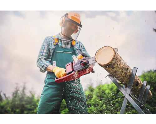 Muž s ochranným oděvem řeže motorovou pilou Einhell kmen stromu na kozlíku