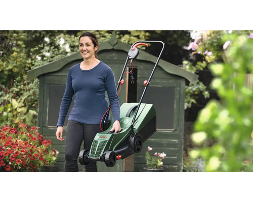 Žena nese elektrickou sekačku na trávu na zahradě před kůlnou na nářadí.