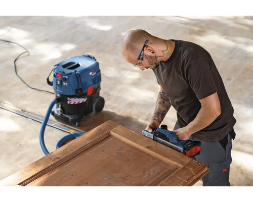 Řemeslník hobluje dřevěné dveře elektrickým hoblíkem a vysavačem