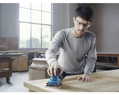 Muž brousí dřevo vibrační bruskou v dílně.