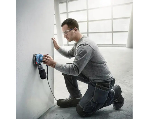 Muž brousí stěnu elektrickou bruskou ve světlé místnosti.