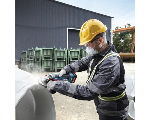 Muž s ochrannou přilbou, ochrannými brýlemi a respirátorem brousí obrobek pomocí akumulátorové úhlové brusky.