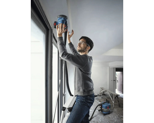 Muž na žebříku brousí strop stropní bruskou, připojen k vysavači