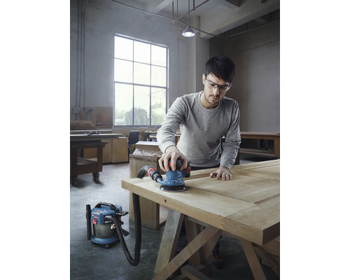 Muž brousí dřevěné dveře pomocí excentrické brusky a vysavače v dílně.