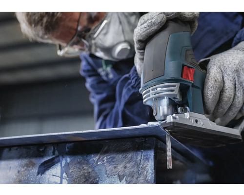 Osoba s respirátorem řeže kus kovu přímočarou pilou.