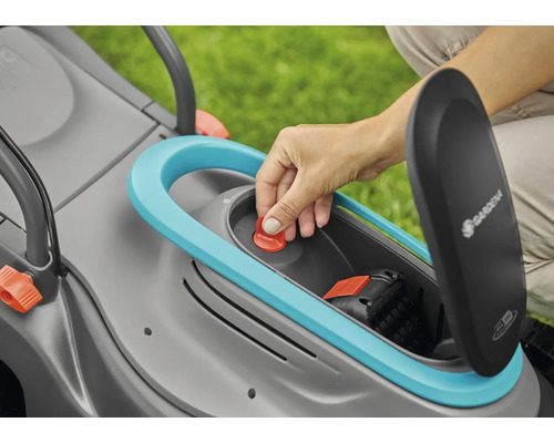 Ruka vkládá červené tlačítko do sekačky na trávu s otevřenou přihrádkou na baterie. Je viditelné logo Gardena.