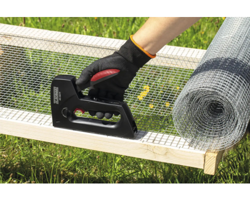 Osoba připevňuje drátěné pletivo sponkovačkou k dřevěnému rámu, s rolí drátěného pletiva v pozadí.