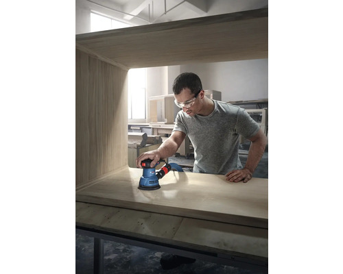 Muž brousí dřevěný kus nábytku excentrickou bruskou značky Bosch.