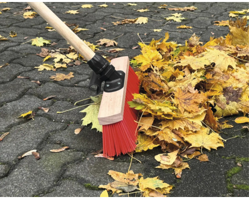 Zametací stroj s dřevěnou násadou a červenými štětinami zametá listí na dlážděné cestě.