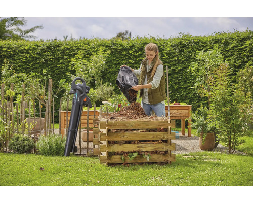 Žena vysypává listí do dřevěného kompostéru na zahradě. Vedle něj stojí foukač listí.