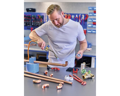 Instalatér pájí měděné trubky hořákem, zatímco na ponku leží různé nástroje a trubkové díly.