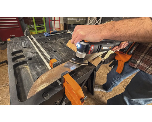 Osoba používá multifunkční nástroj k broušení obrobku, který je připevněn svorkami k pracovnímu stolu.