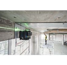 Zelený čárový laser je připevněn k betonovému nosníku a promítá zelenou laserovou čáru. V pozadí stojí pracovník na lešení.