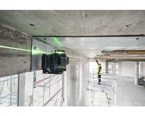Zelený čárový laser je připevněn k betonovému nosníku a promítá zelenou laserovou čáru. V pozadí stojí pracovník na lešení.