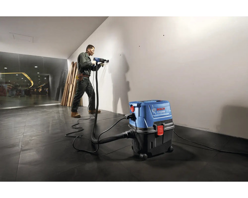 Řemeslník vrtá vrtačkou do zdi, zatímco průmyslový vysavač odsává prach.