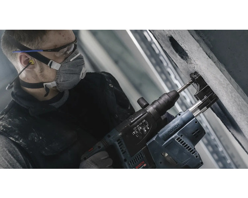 Dělník vrtá s vrtacím kladivem otvor do betonové zdi. Má na sobě ochrannou masku, ochranné brýle a chrániče sluchu.