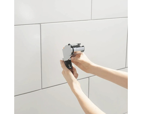 Ruce drží držák sprchové hlavice před obloženou stěnou.