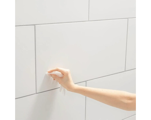 Ruka čistí bílou obkládanou stěnu hadrem.