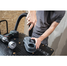 Řemeslník připevňuje hubici vysavače k odsávání prachu z vrtání na pracovní lavici. Vedle leží vrtačka a děrovací pilový list.