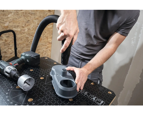 Řemeslník připevňuje hubici vysavače k odsávání prachu z vrtání na pracovní lavici. Vedle leží vrtačka a děrovací pilový list.