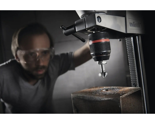 Stojanová vrtačka s mužem s ochrannými brýlemi, který vrtá otvor do kusu kovu.