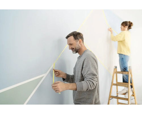 Dvě osoby zdobí stěnu maskovací páskou a geometrickými tvary.
