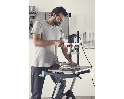 Muž s ochrannými brýlemi vrtá vrtačkou do obrobku na pracovním stole.