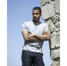 Mladý muž se zapletenými vlasy se opírá o kamennou zeď se založenýma rukama, má na sobě tričko a kapsáčové kalhoty.