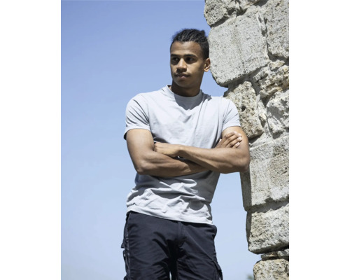 Muž se zkříženýma rukama opřený o kamennou zeď, oblečený do trička a cargo šortek