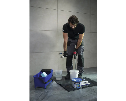 Řemeslník míchá lepidlo na dlaždice s míchadlem barev před obloženou stěnou. Modrý čisticí kbelík a nádoba s lepidlem jsou připraveny.