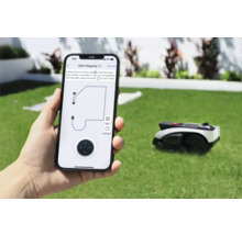 Osoba drží smartphone s aplikací pro ovládání robotické sekačky na trávu. V pozadí je robotická sekačka na trávníku.