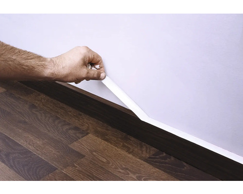 Upevnění bílé podlahové lišty na stěnu nad tmavou dřevěnou podlahou.