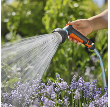 Ruka drží zahradní hadici se sprchovou hlavicí, která stříká vodu na květiny.