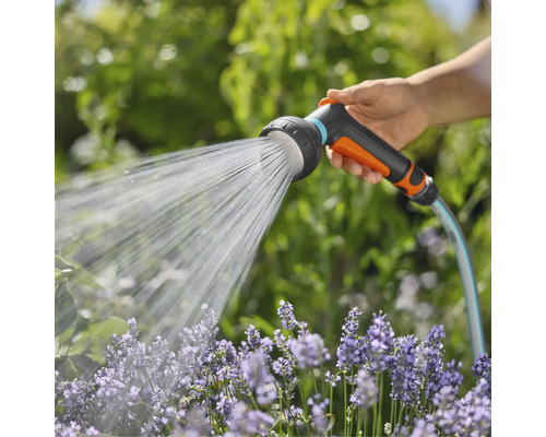 Ruka drží zahradní hadici se sprchovou hlavicí, která stříká vodu na květiny.