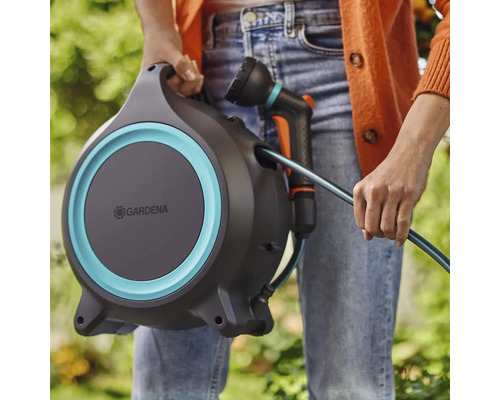 Osoba drží hadicový box Gardena se zahradní hadicí a postřikovačem.