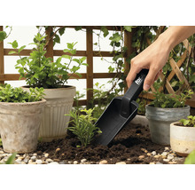 Zahradní scéna s květináči, okolní zelení a osobou používající sázecí lopatku.