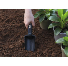 Ruka drží sázecí lopatku v zemi, vedle hrnkových rostlin.