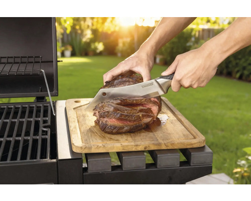 Připravený steak se krájí nožem na dřevěném prkénku vedle grilu.