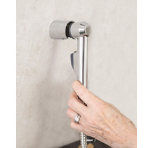 Ruka drží bidetovou sprchu se sprchovou hadicí na nástěnném držáku v koupelně.