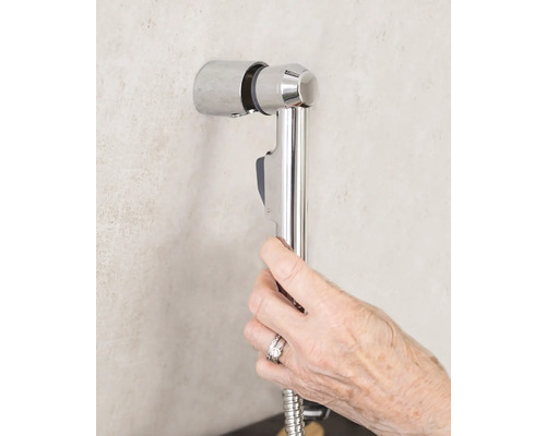 Ruka drží bidetovou sprchu se sprchovou hadicí na nástěnném držáku v koupelně.