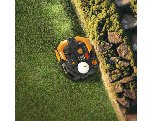 Pohled shora na robotickou sekačku na trávě vedle kamenné zdi.