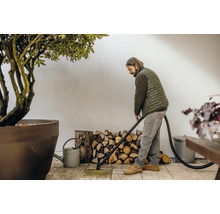 Muž čistí terasu vysavačem popela, v pozadí hromada dřeva a rostlina