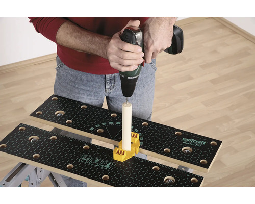 Osoba vrtá vrtačkou do kusu dřeva, který je upevněn na pracovním stole se žlutým upevňovacím prvkem.