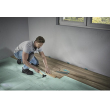 Muž pokládá laminátovou podlahu s kladivem a tloukem na kročejovou izolaci v místnosti.